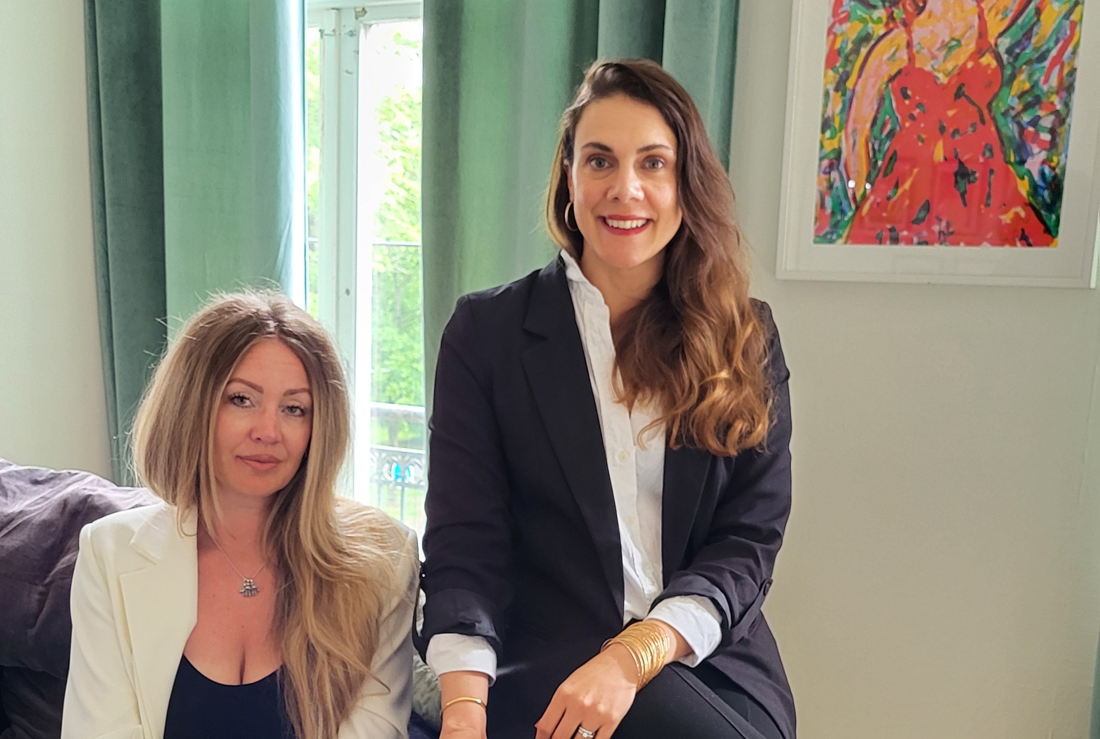 En blond kvinna i vit kostym sitter i en soffa och på armstödet sitter en mörkhårig kvinna i svart kostym . Text: För livet - tillsammans med Johanna och Maria.